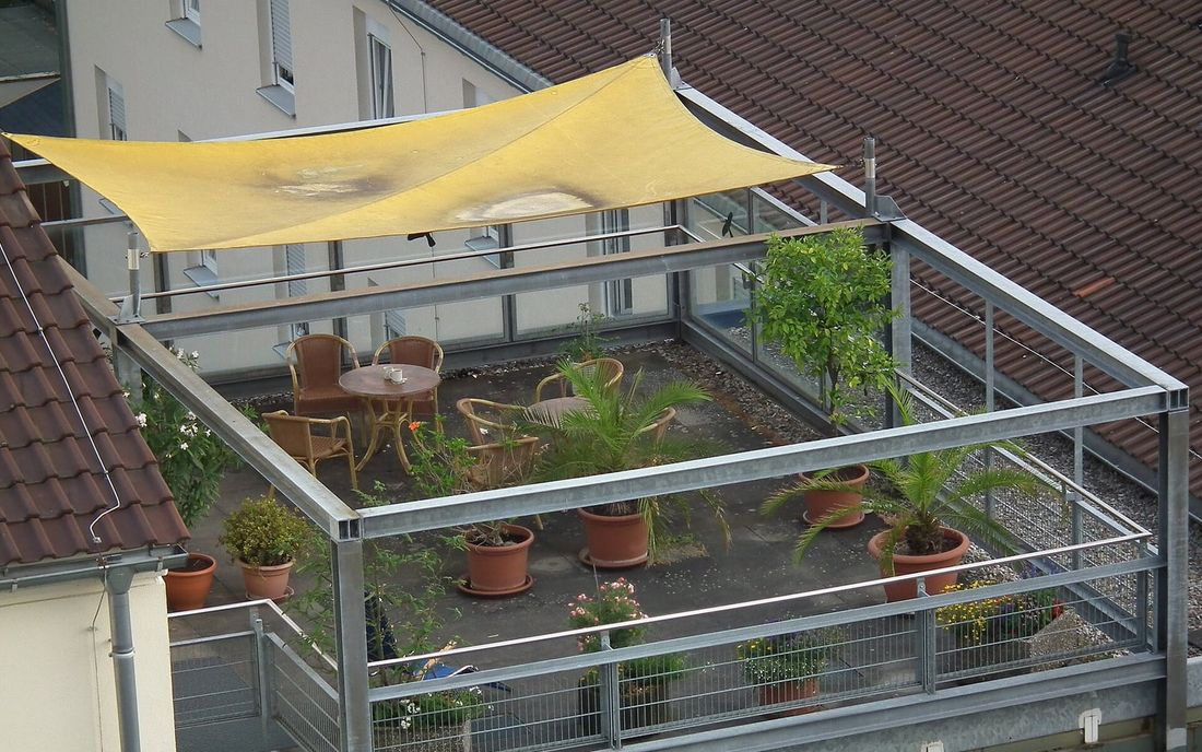 Terraço com guarda-corpo metálico, mesa, cadeiras e vasos de plantas sob uma vela de sombreamento amarela